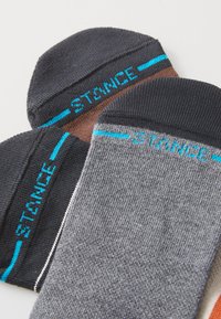 Dos pares de calcetines: uno negro con acentos azules y otro gris con detalles en azul y marrón. Material de punto, acabado texturizado y logo visible.