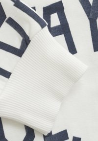 Tela blanca con texto en azul marino, con un puño acanalado. La tela es suave con un acabado texturizado. El diseño incluye letras grandes.