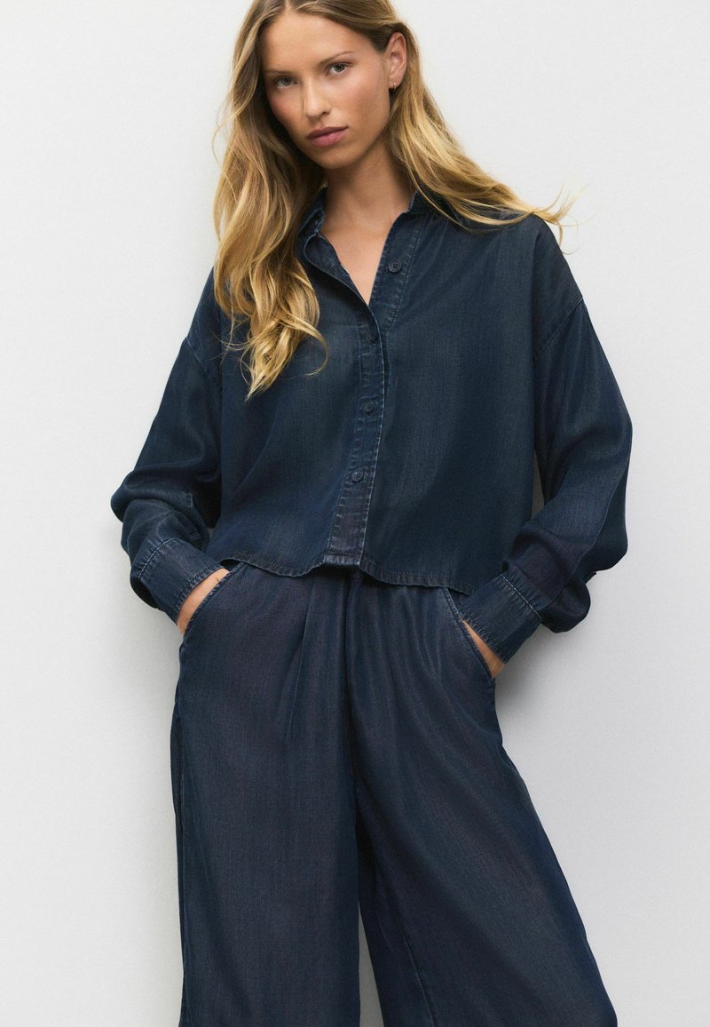 Chemise en denim bleu foncé à manches longues, avec un devant boutonné et des poches. Associée à un pantalon taille haute ample assorti. Texture douce.