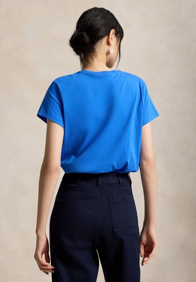 Frau mit dunklen, zu einem Dutt gebundenen Haaren, trägt ein hellblaues kurzärmeliges Hemd und schwarze Hose, von hinten vor neutralem Hintergrund gezeigt.