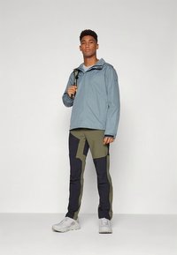 Ung mand iført en lyseblå jakke, olivengrønne og sorte bukser, hvide sneakers, holder en rygsækrem over den ene skulder.