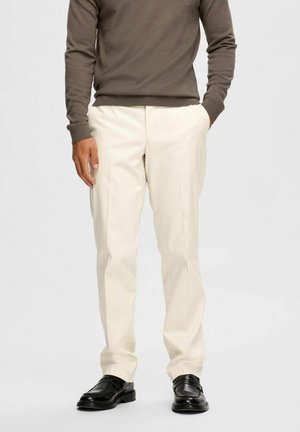 Cremefarvede bukser med skræddersyet pasform, med sidelommer og en glat tekstur, parret med sorte loafers og en brun sweater.
