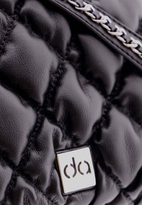 Bolso de cuero negro acolchado con una etiqueta de logotipo cuadrado plateado brillante y un detalle de cadena metálica. La textura parece suave y acolchada.