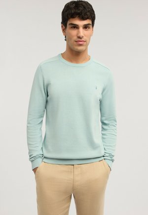 Hombre con cabello rizado oscuro, vestido con un suéter azul claro y pantalones beige, de pie con las manos en los bolsillos frente a un fondo liso.