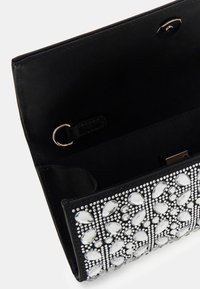 Pochette nera aperta che mostra l'interno, decorata con grandi e piccoli gioielli trasparenti disposti in un motivo floreale sulla patta.