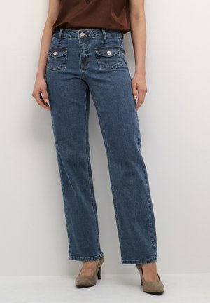Femme portant un jean bleu taille haute avec des poches à rabat boutonnées, associé à des chaussures à talons marron et un top marron sans manches.