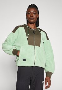 Lys grøn fleece hettejakke med brune paneler, glidelås og frontlommer. Har snor og et merke nær kanten.