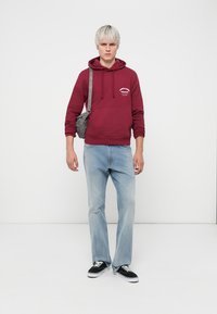 Hoodie en coton bordeaux avec poche avant et graphique blanc. Jean droit bleu clair. Baskets noires avec semelles blanches. Sac bandoulière gris.