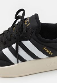 Juodos sportinės bateliai su baltomis zigzago juostelėmis, auksiniu "SAMBA" tekstu, apvaliu nosiu, tekstūruotu audiniu viršuje ir tvirtu smėlio spalvos guminėmis padangomis.