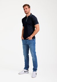 Námořnická modrá polo košile s bílým zipem u límce, nošena s světle modrými džíny a bílými teniskami s černým logem. Hladký materiál, přiléhavý střih.