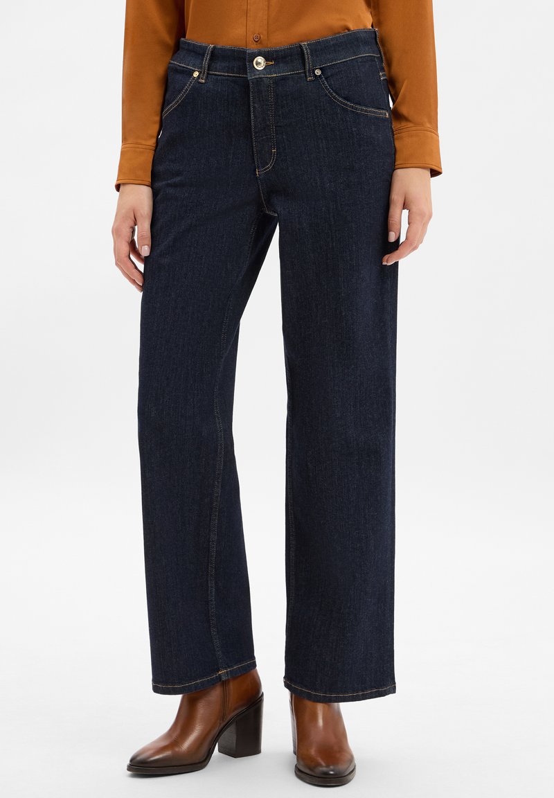 Jean en denim foncé avec un design à jambes droites, dotés de surpiqûres dorées, d'une fermeture à bouton et à glissière, ainsi que de poches avant. Associé à des bottes à talons marron.