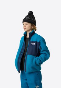 Μπλε φλις μπουφάν με ναυτικές λεπτομέρειες, πλήρης φερμουάρ, δύο μπροστινές τσέπες. Μαύρο πλεκτό σκουφάκι με σχέδιο και φούντα. Μαύρο πουκάμισο από κάτω.