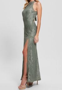 Olivengrøn maxikjole med geometriske udskæringer, høj sideløsning og halterneck. Har lagdelt tekstur og indviklede syningsdetaljer.