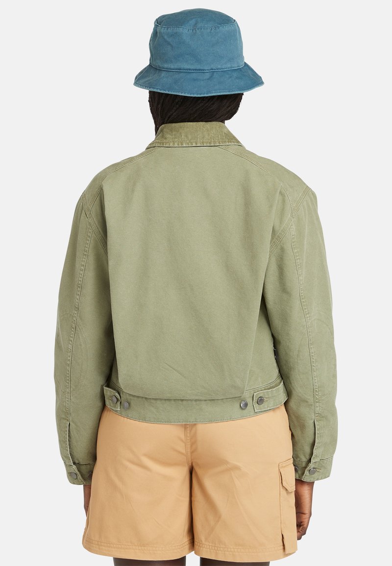 Persona che indossa una giacca verde chiaro e pantaloni cargo beige, con cappello a secchiello blu, vista da dietro su uno sfondo bianco uniforme.