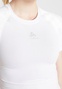 Bílá sportovní košile Odlo se střihovým designem, texturovanou látkou s jemnými síťovanými vzory a nenápadným logem na hrudi.