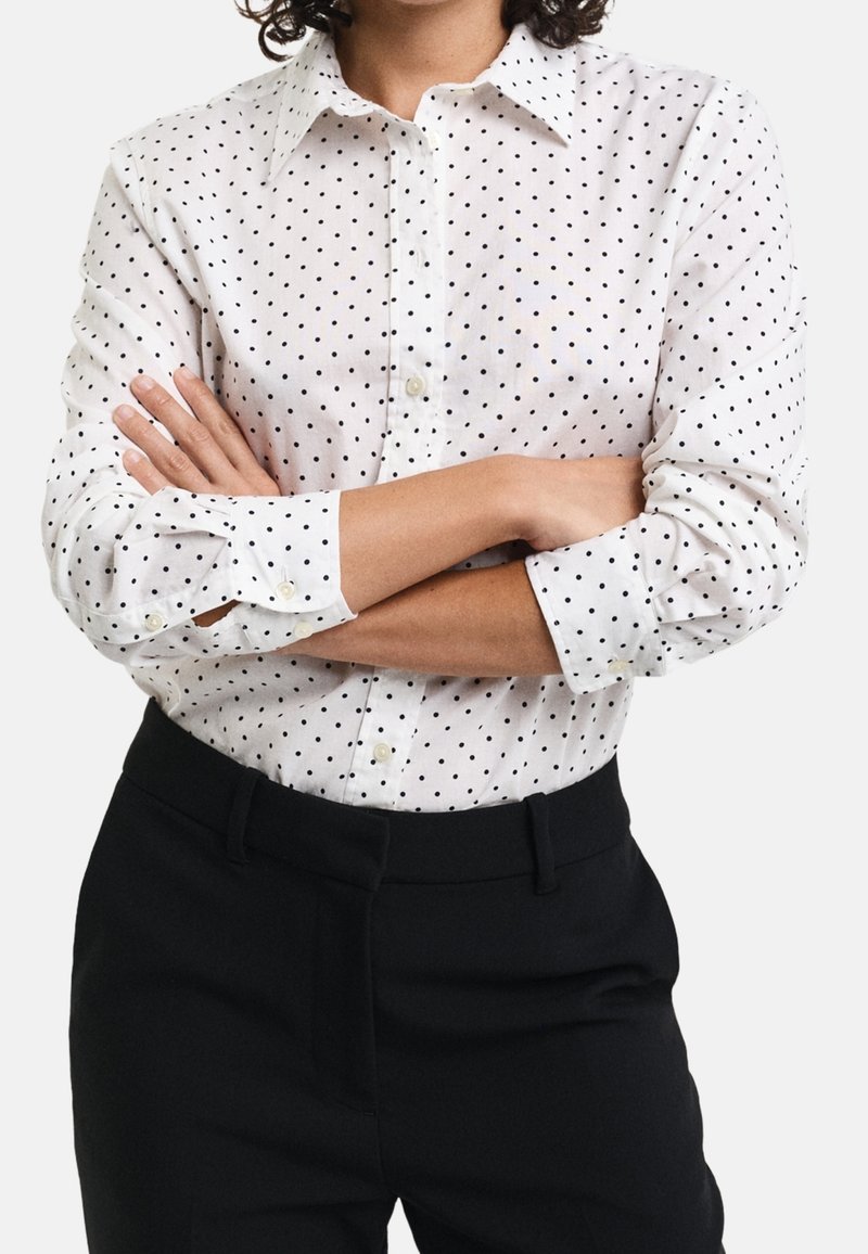 Personne portant une chemise blanche à boutons avec des pois noirs, les bras croisés sur un pantalon noir, sur un fond uni.