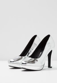 Para błyszczących srebrnych szpilek z platformą, czarnymi wkładkami i szpilkowymi obcasami na białej powierzchni.