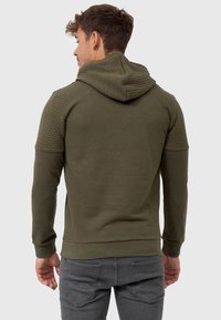 Olijfgroene hoodie met textuur en gewatteerde schouderaccenten, lange mouwen en een aansluitende zoom. De stof lijkt zacht en rekbaar.