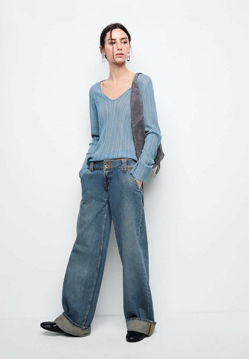 Femme portant un jean bleu large, un pull côtelé bleu clair, des chaussures noires et un sac à épaule gris, debout contre un mur blanc.