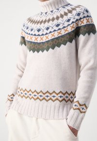 Sweter z golfem wykonany z dzianiny w jasnym kremowym kolorze, z geometrycznymi wzorami w odcieniach niebieskiego, zielonego, pomarańczowego i brązowego na ramionach i u dołu.