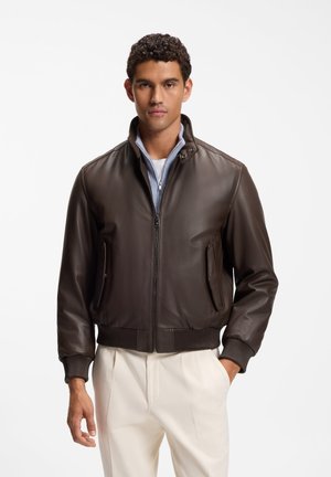 Man draagt een donkerbruine leren jas over een lichtblauw shirt met rits en crèmekleurige broek, staand met één hand in zijn zak.