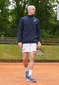 Sportos férfi tengerészkék zipzáras dzsekiben és fehér sortban, teniszkészlettel a kezében, kék sportcipőben, fehér részletekkel. Szabadtéri teniszpálya.