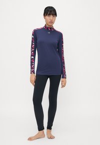 Marineblauer Rash Guard mit langen Ärmeln und gepunkteten rosa und weißen Ärmeln, ausgestattet mit einem hohen Kragen. Kombiniert mit schwarzen Leggings mit strukturierten Bündchen.