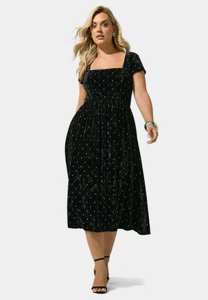 Femme portant une robe midi en velours noir à pois argentés, encolure carrée, manches courtes, escarpins noirs à bride cheville et un bracelet argenté.