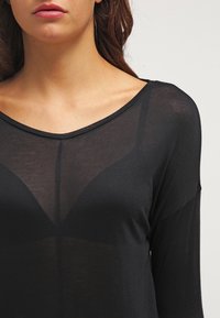 Žena v průhledném černém topu přes černou podprsenku, ukazující klíční kosti a část rtů a brady na jednobarevném pozadí.