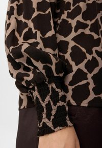 Maglietta a maniche lunghe con stampa giraffa marrone e nera, con polsino elastico arricciato, appoggiata sul braccio di una persona su uno sfondo neutro.
