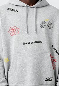 Felpa grigia con cappuccio e ricami colorati assortiti, tra cui una rosa rossa, testo nero, una stella gialla e un dettaglio di un angelo con ali.