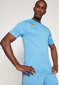 Camisola atlética de um azul claro, feita de tecido suave, com mangas curtas e decote redondo. Acentos de logótipo dourado nos ombros.