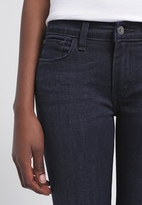 Jeans de denim azul oscuro con un diseño ajustado, estilo de cinco bolsillos y un botón plateado visible en la cintura. Textura suave con ligero desgaste.