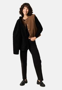 Ruskotäytteinen ruskea ja musta liivi napilla, which is worn over a black long-sleeve top, associated with black jeans and shoes.