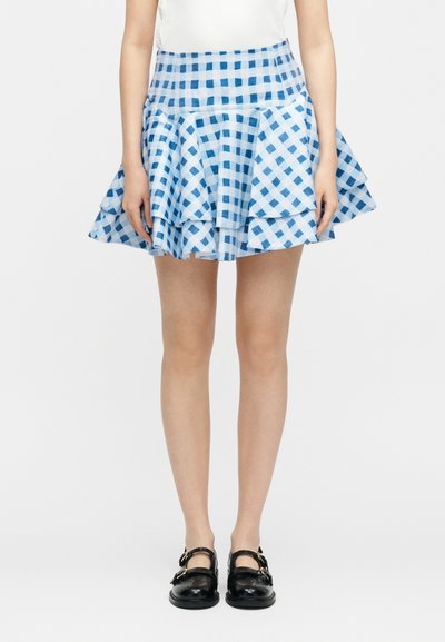 Γυναίκα με μπλε και άσπρη μίνι καρό φούστα με βολάν, συνδυασμένη με μαύρα παπούτσια Mary Jane, στέκεται μπροστά σε ένα απλό λευκό φόντο.