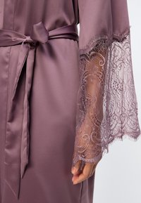 Μωβ σατέν ρόμπα με δεμένη ζώνη στη μέση και διάφανα δαντελωτά μανίκια σε σχήμα καμπάνας που αποκαλύπτουν μερικώς ένα χέρι κάτω από τις δαντέλες.