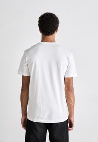 Camiseta de algodón blanca de manga corta, con cuello redondo y una textura suave, presentada desde la parte de atrás. Combinada con pantalones oscuros.
