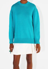 Turkusowy sweter dziergany z okrągłym dekoltem i ściągaczami, charakteryzujący się luźnym krojem i bocznymi rozcięciami. Nosi się go na plisowanej białej spódnicy.