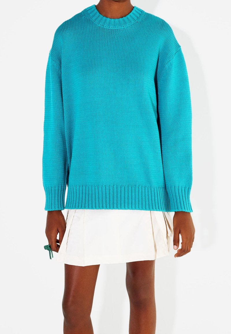 Turkusowy sweter dziergany z okrągłym dekoltem i ściągaczami, charakteryzujący się luźnym krojem i bocznymi rozcięciami. Nosi się go na plisowanej białej spódnicy.