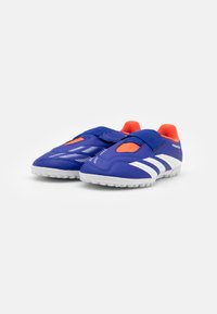 Zapatillas deportivas azules con un exterior liso, presentan un diseño de tres rayas blancas, detalles en naranja y una correa de Velcro para un ajuste seguro.