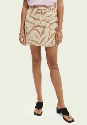 Mujer vistiendo una minifalda beige y marrón con estampado, cremallera frontal y bolsillos, combinada con sandalias negras de tacón y puntera abierta.