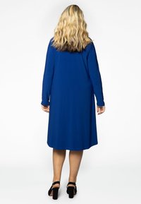 Blaues Kleid mit langen Ärmeln und lockerem Schnitt, knielanger Saum und glattem Stoff. Getragen mit schwarzen High-Heels.
