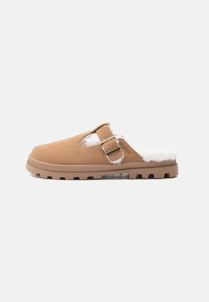 Beige Wildleder Hausschuh mit weißem Kunstfellfutter, verstellbarem Riemen und einer strukturierten Gummisohle mit kleinen Traktionselementen.
