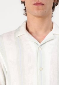 Camicia button-up realizzata in tessuto leggero, con una texture morbida e righe verticali nei toni di bianco, verde e azzurro chiaro.