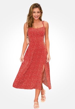 Kobieta uśmiechająca się, ubrana w czerwoną, bezrękawną sukienkę midi w białe kropki z bocznym rozcięciem, beżowe sandały na paski oraz niosąca małą beżową torebkę na ramię.