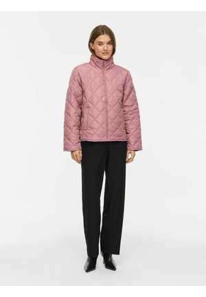 Femme portant une veste matelassée rose à col haut, un pantalon large noir et des bottes noires à bout pointu, debout devant un fond blanc uni.