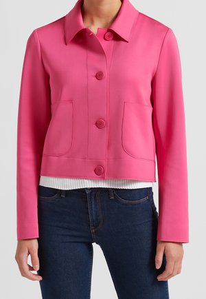 Vrouw draagt een felroze jas met knopen en twee voorzakken over een wit geribbelde top en donkerblauwe spijkerbroek.