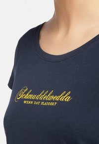 Tummansininen puuvillat-paita, jossa on pyöreä pääntie ja etupuolella keltaista käsinkirjoitettua tekstiä "Schmuddelwedda" ja "Wenn dat pladdert".