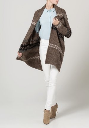 Femme portant un cardigan marron à motifs par-dessus une chemise bleu clair boutonnée, un pantalon blanc, et des bottines en daim beige, posant devant un fond uni.