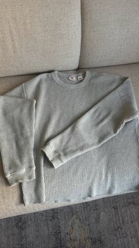 Pilkas briaunotas megztinis, padėtas plokščiai, turi ilgas rankoves ir apvalų iškirpimą. Auditorija yra vienoda, su subtiliu tempimu ir švelniu audinio paviršiumi.